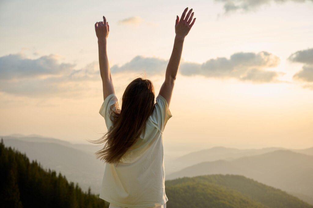 a woman stands on a mountain ridge with arms raised high celebrating the beauty of nature as the sun sets in the background