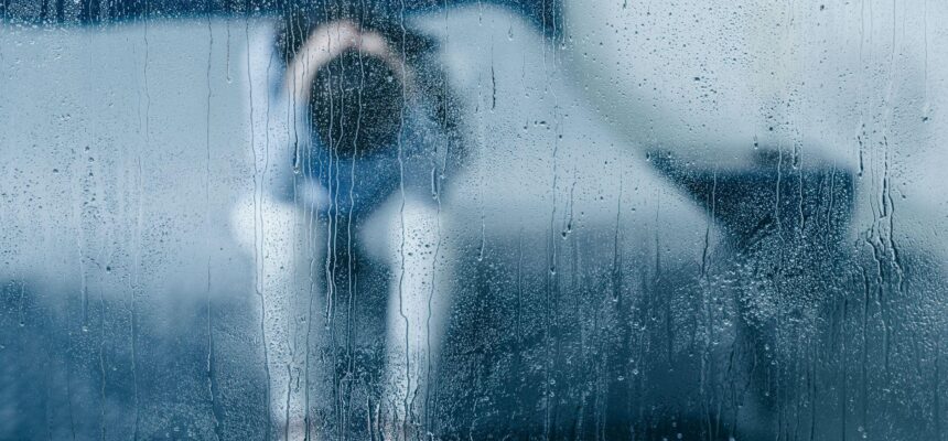depressed woman sitting on bed and holding head in hands through window with raindrops