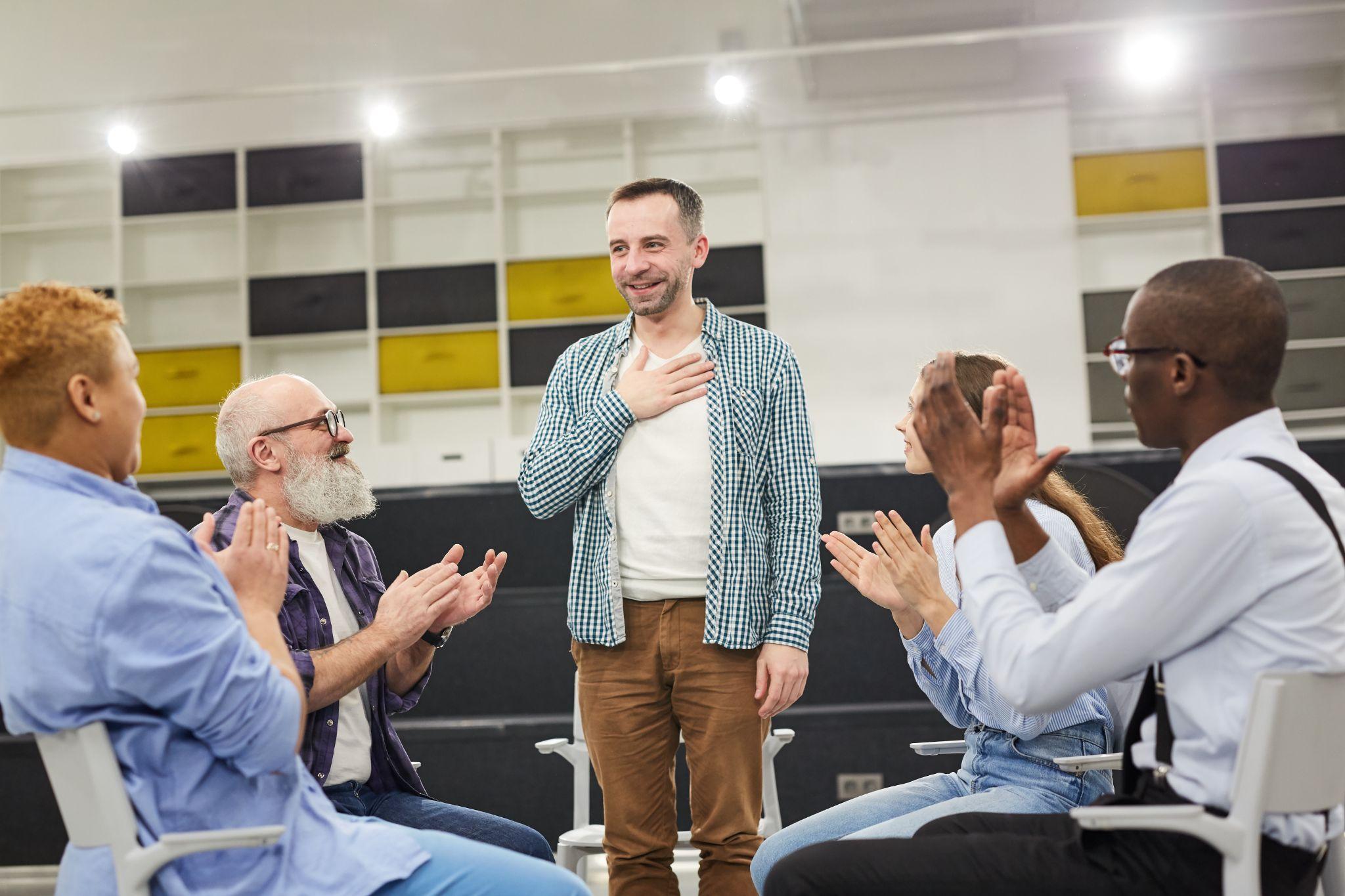 Portrait of smiling mature man introducing himself during therapy session in support group to people clapping