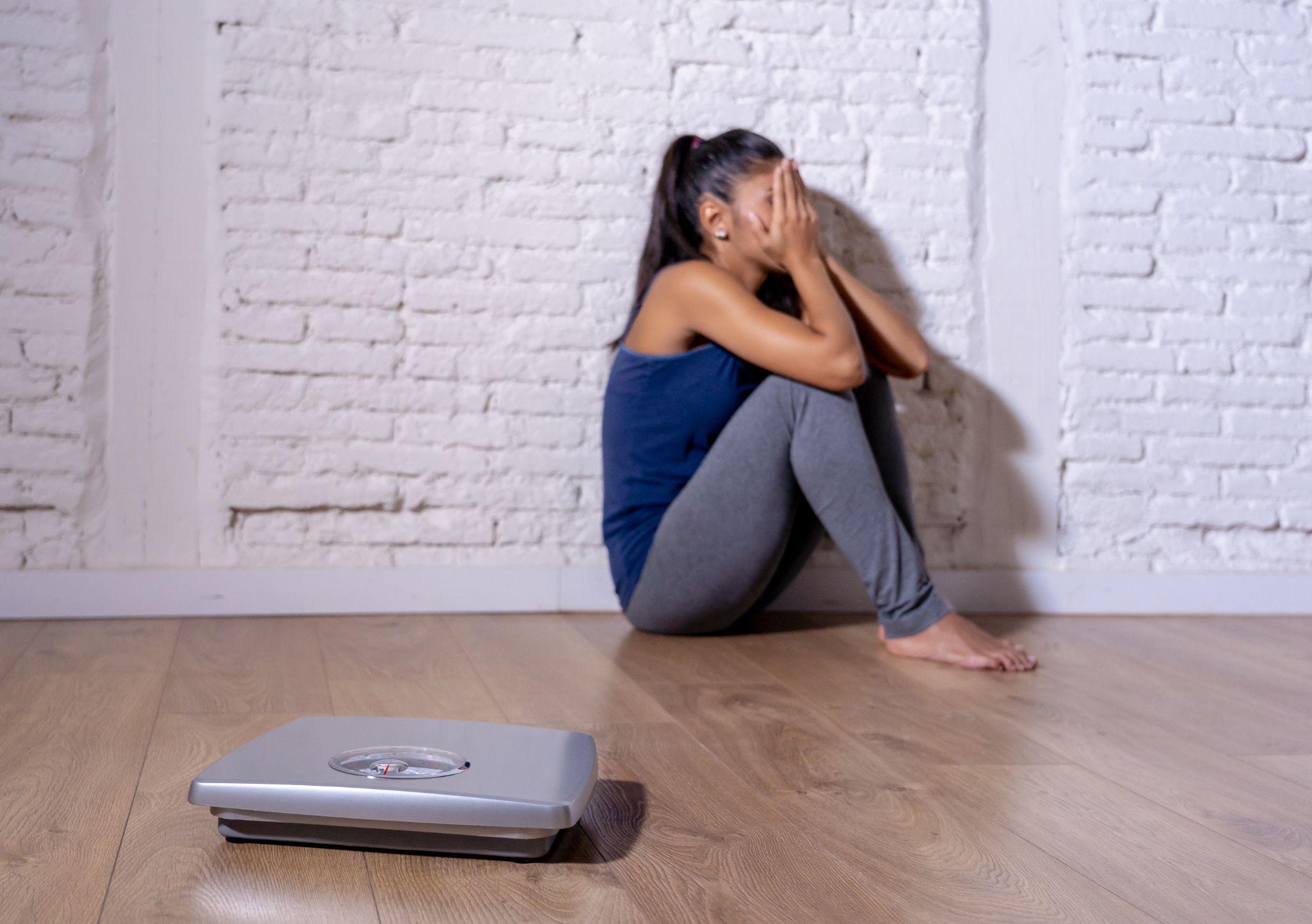 Young teenager woman sitting alone on ground looking at the scale worried and depressed in dieting and eating disorder